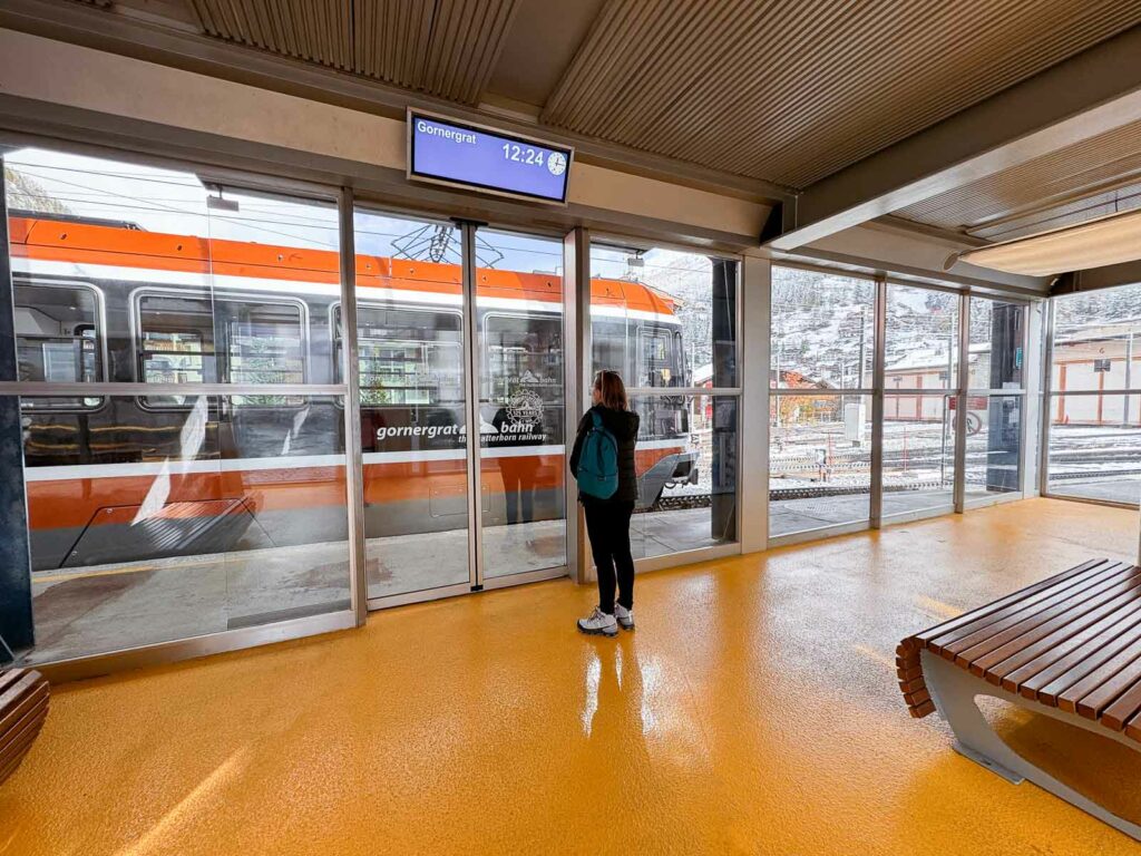 A traveller stands in the priority waiting area of the Gornergratbahn, with the iconic orange and white train visible through the glass, ready to discover the best side to sit on the Gornergrat train for panoramic views