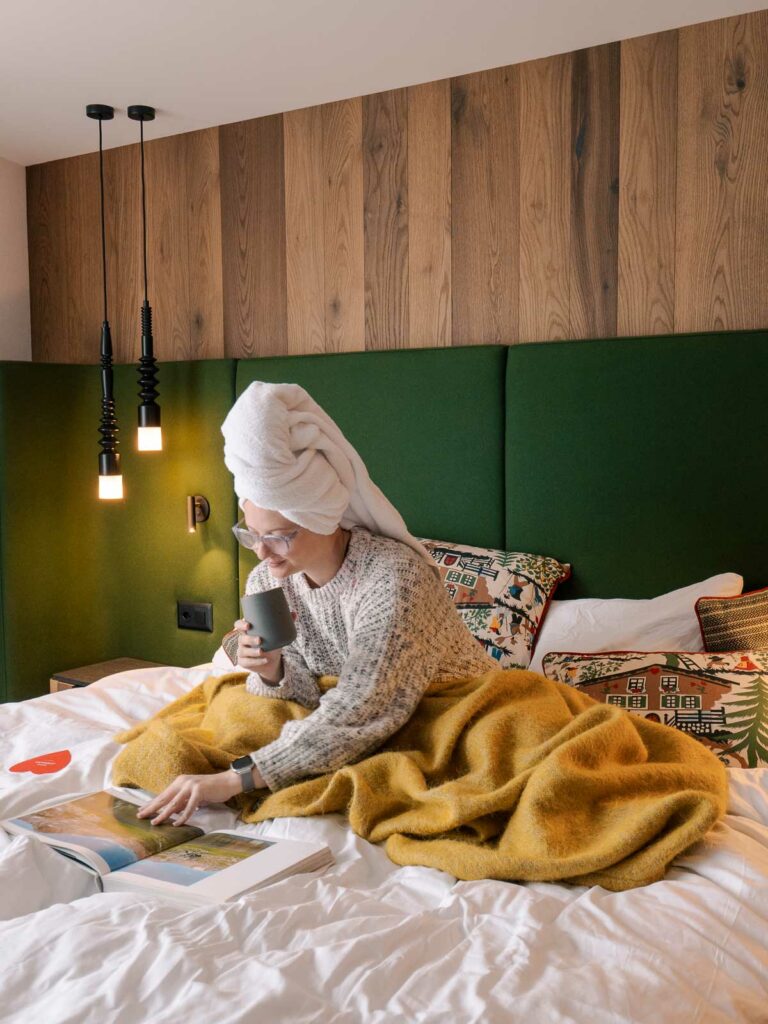 Alexx sits on a bed at Hotel Zermama, the best hotel in Zermatt, engrossed in a travel guide, with a modern wooden headboard and stylish pendant lights in the background
