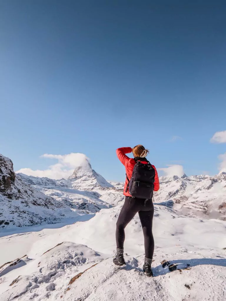 A hiker in a red jacket stands against the snowy landscape near Rotenboden station, looking towards the majestic Matterhorn, embodying the spirit of adventure in the Swiss Alps
