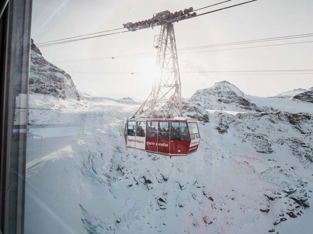 A red cable car makes its way up to Matterhorn Glacier Paradise against a backdrop of rugged, snow-covered slopes, seen through the window of another cable car, showcasing a unique perspective of the ascent during a ten day Switzerland itinerary