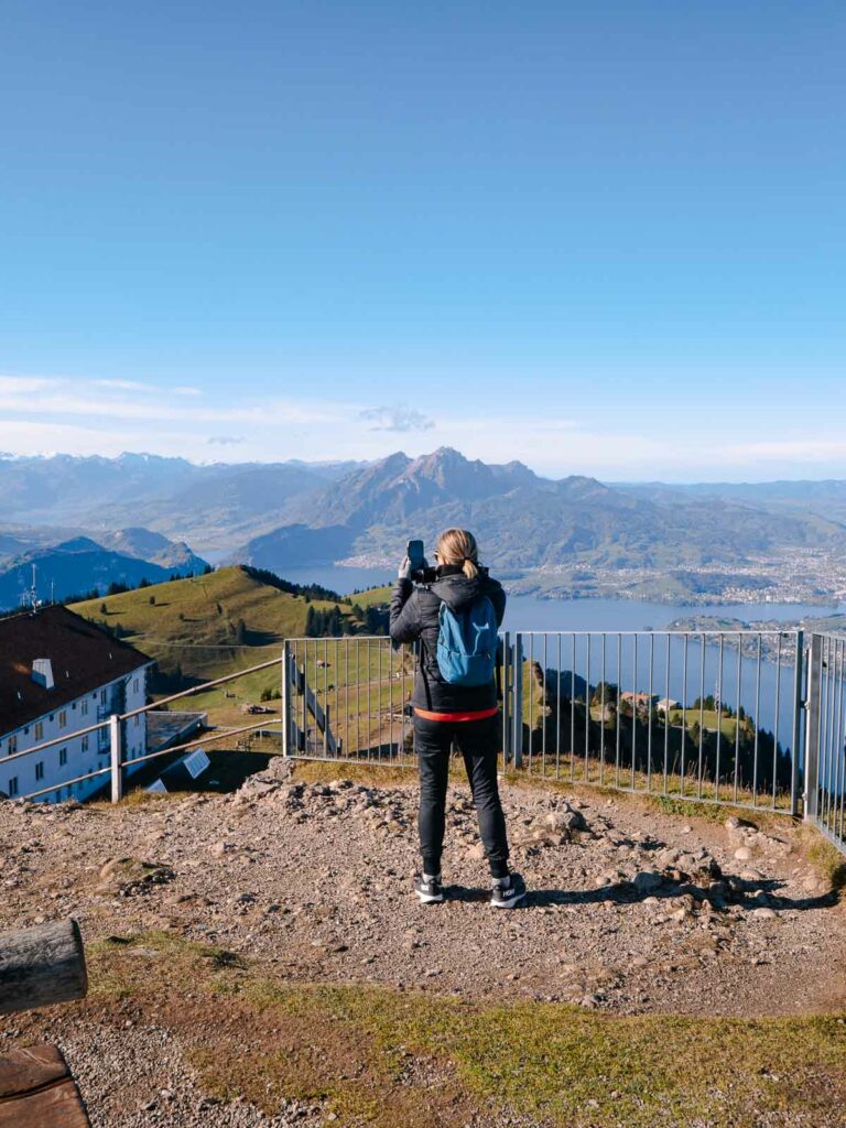 A traveller captures a moment atop Mount Rigi on their Switzerland itinerary, with panoramic views of the surrounding Alps and Lake Lucerne in the distance, beneath the expansive blue sky