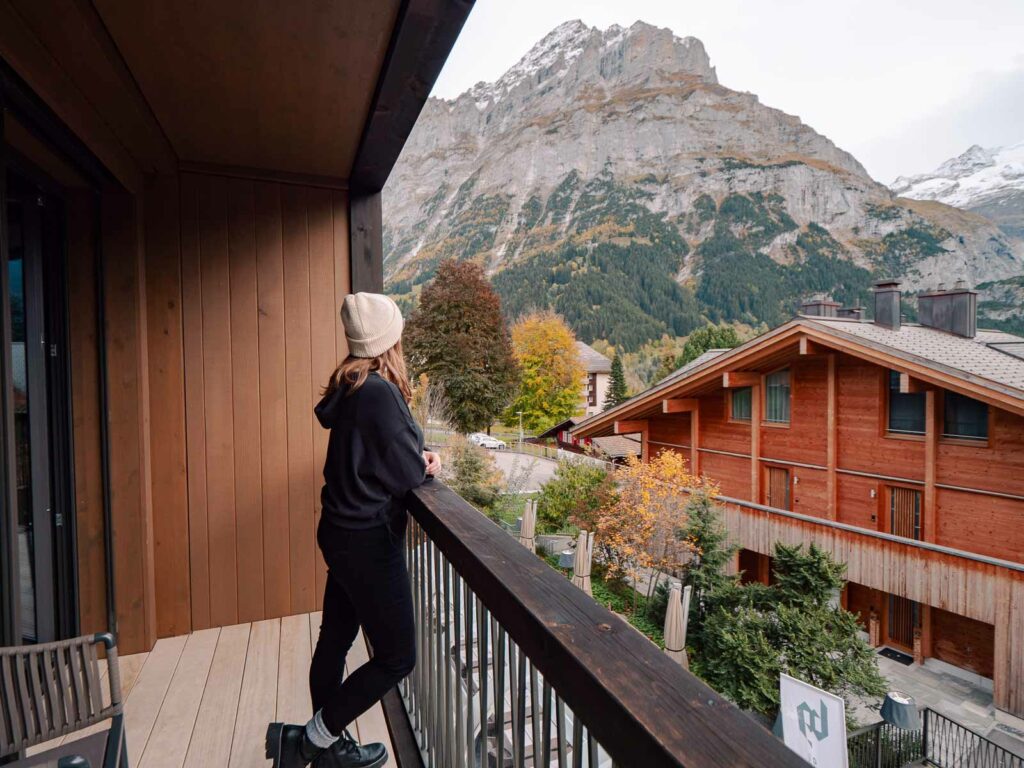 Alexx enjoys the view from the balcony of Bergwelt Design Hotel, overlooking Swiss chalets and the rugged peaks of the Alps, capturing the essence of a tranquil Swiss getaway