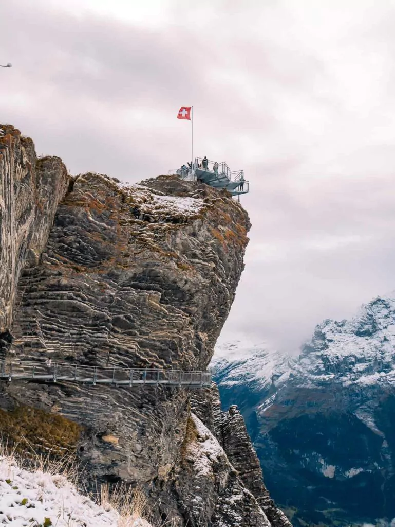 A rugged cliff adorned with a Swiss flag stands prominently at Grindelwald First, with a cliff walk wrapping around it, providing a dramatic view over the alpine landscape
