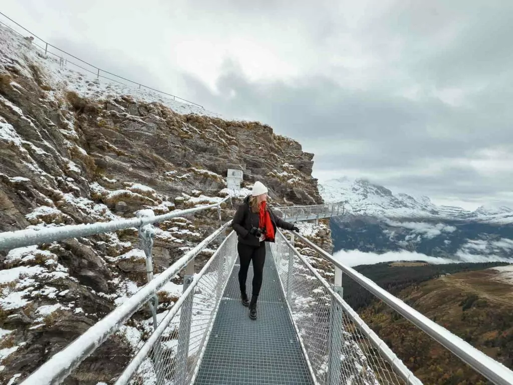 person with a red backpack walks along the Cliff Walk at Grindelwald-First, with dramatic mountain scenery and overcast skies in the background