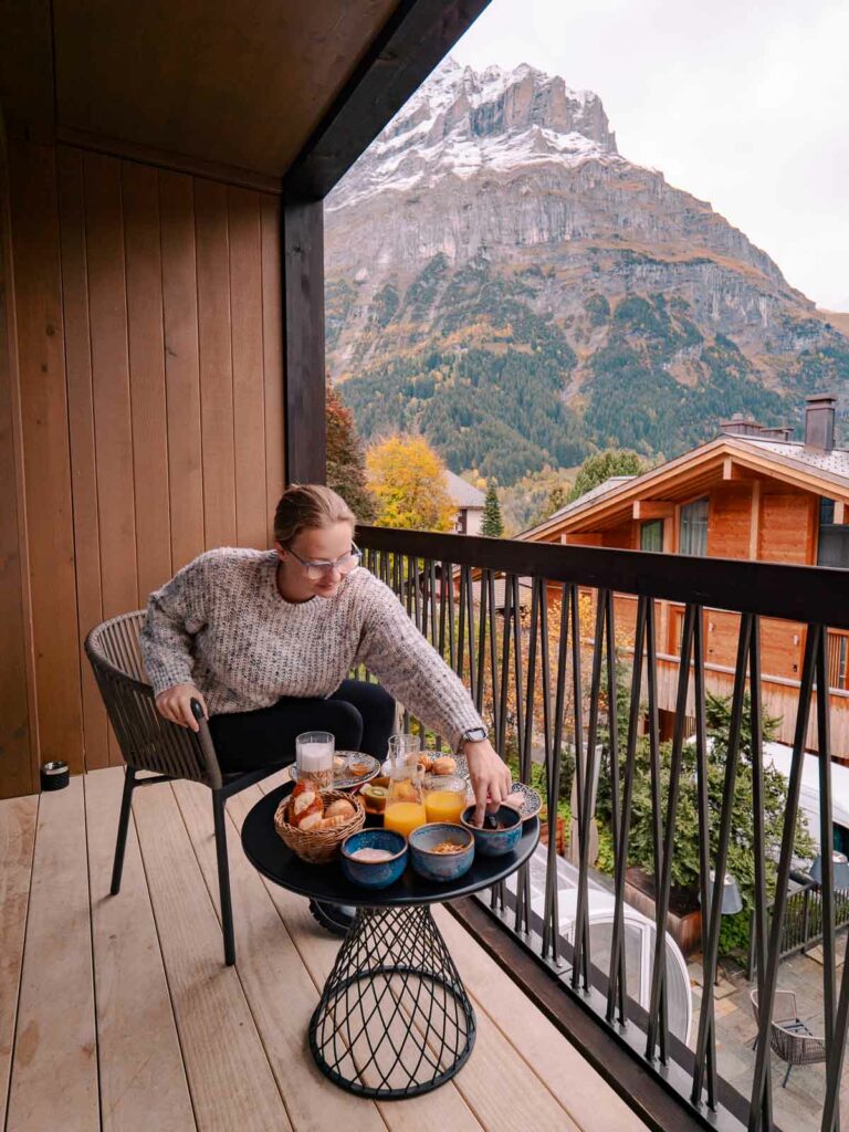 A person relaxes on the balcony of Bergwelt Design Hotel, enjoying a breakfast spread with a stunning view of the mountainous landscape in Grindelwald during a Switzerland adventure