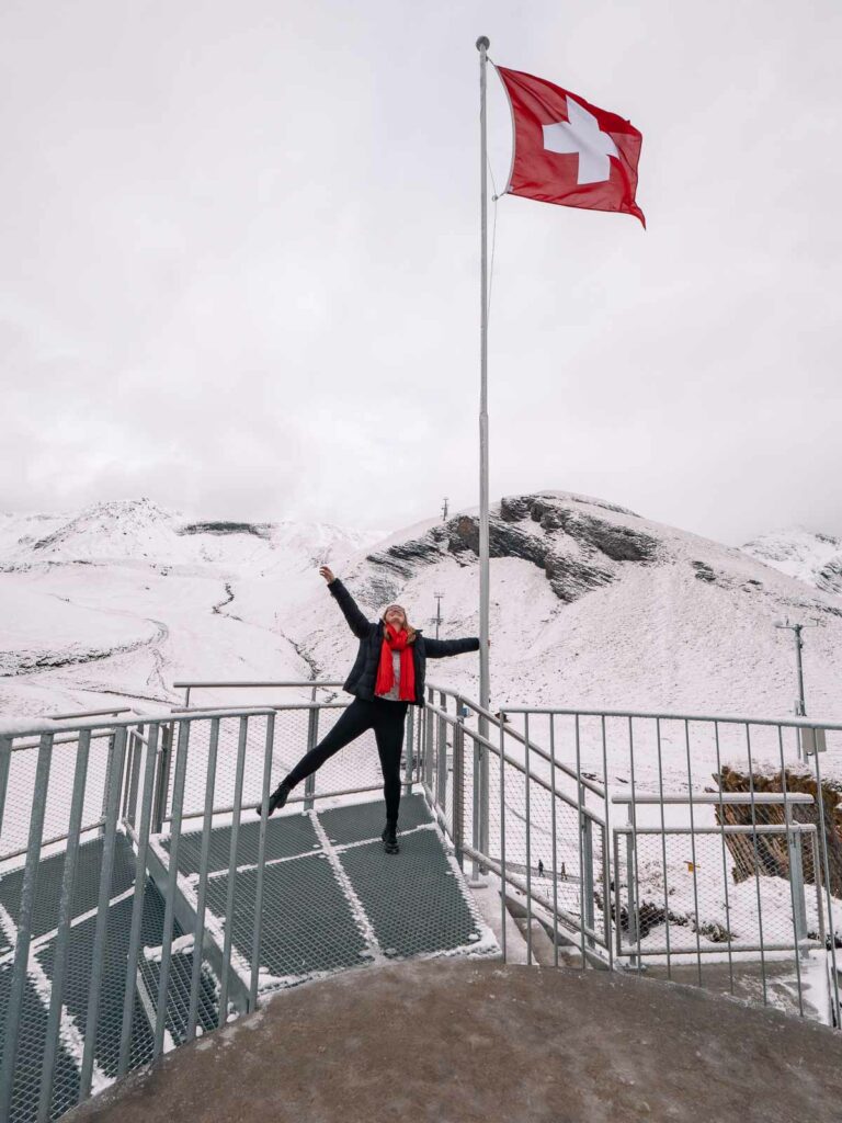 A joyful individual strikes a playful pose at the summit of Grindelwald-First, with a Swiss flag fluttering in the wind and a backdrop of snow-covered mountain peaks