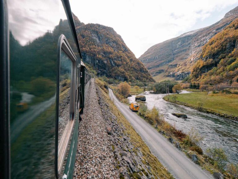 A serene view from the Flam to Myrdal train, with a mirror-like river running parallel to the track, flanked by the gentle slopes of Norwegian mountains in autumn
