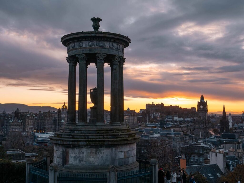 The Dugald Stewart Monument on Calton Hill silhouetted against a dramatic sunset sky, with Edinburgh’s city lights beginning to twinkle