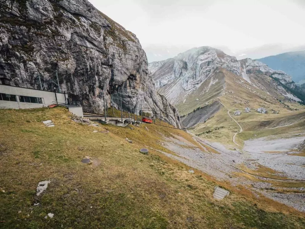 Lush, green valleys descending from the rocky, alpine peak of Mount Pilatus, with a small, visible cogwheel train ascending along the mountainside against a backdrop of distant, mist-shrouded mountains under a dramatic sky