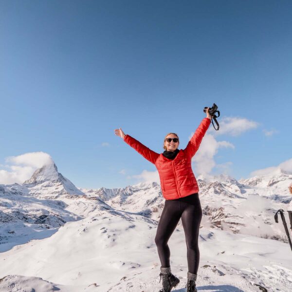 Alexx in a red jacket and sunglasses raising their arms in joy, standing in the snow at the Rotenboden station on the Gornergrat railway line with the Matterhorn in the background