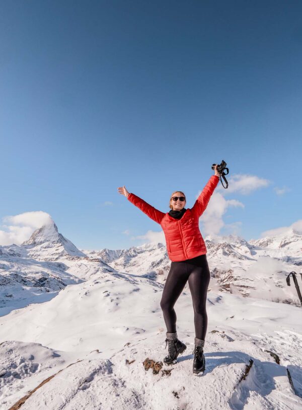 Alexx in a red jacket and sunglasses raising their arms in joy, standing in the snow at the Rotenboden station on the Gornergrat railway line with the Matterhorn in the background