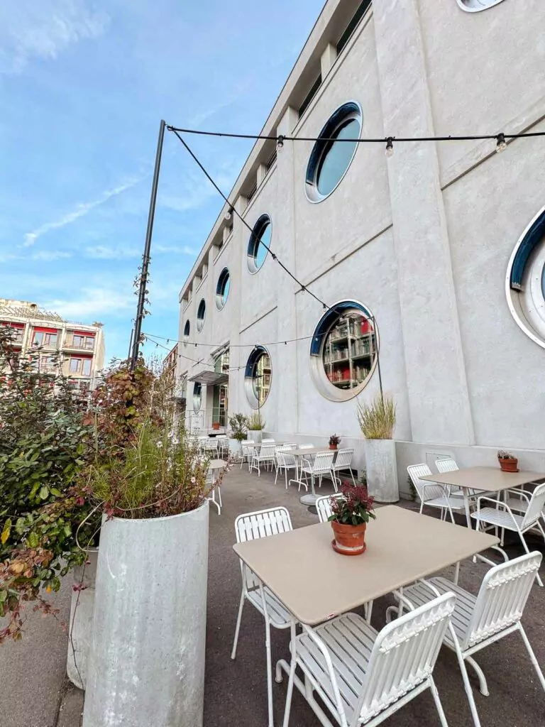 An outdoor seating area with white metal tables and chairs set against the exterior of Silo Hostel Basel, characterised by its large, round porthole windows and strung lights above