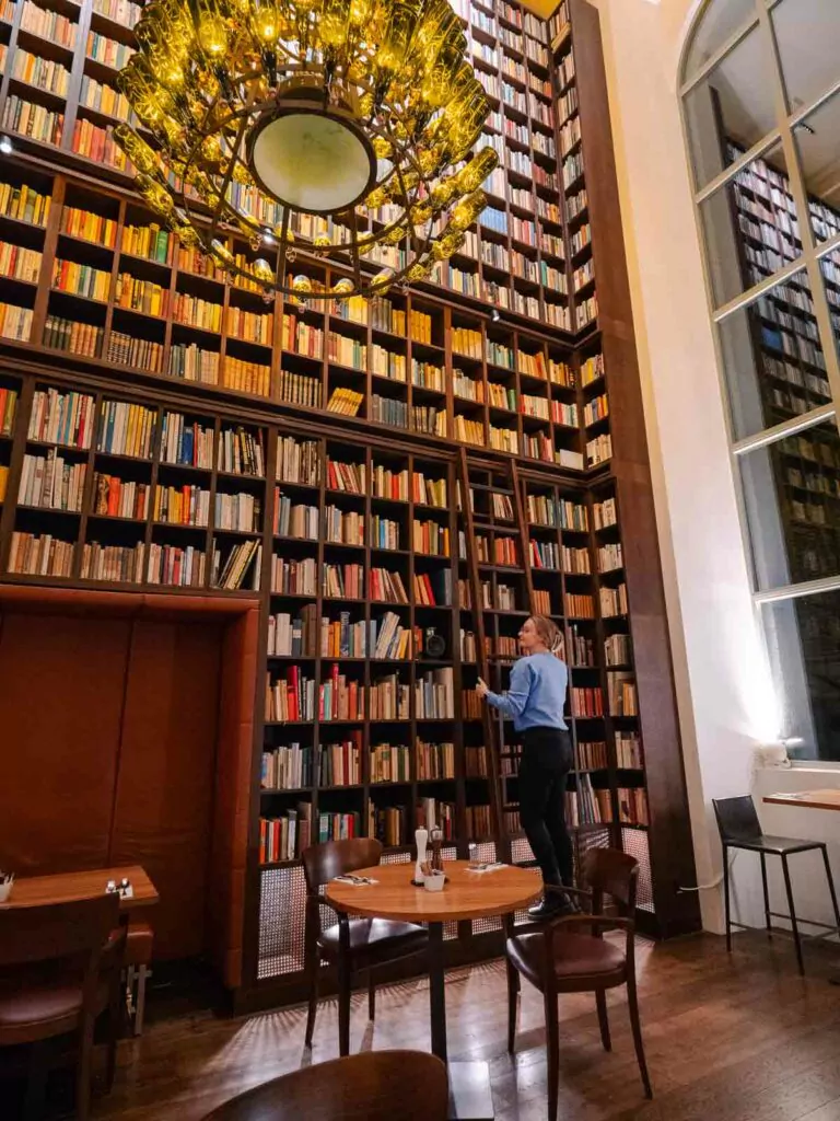 Interior of the B2 Hotel library in Zurich with Alexx reading a book by tall bookshelves under a decorative chandelier, beside large arched windows and wood furnishing