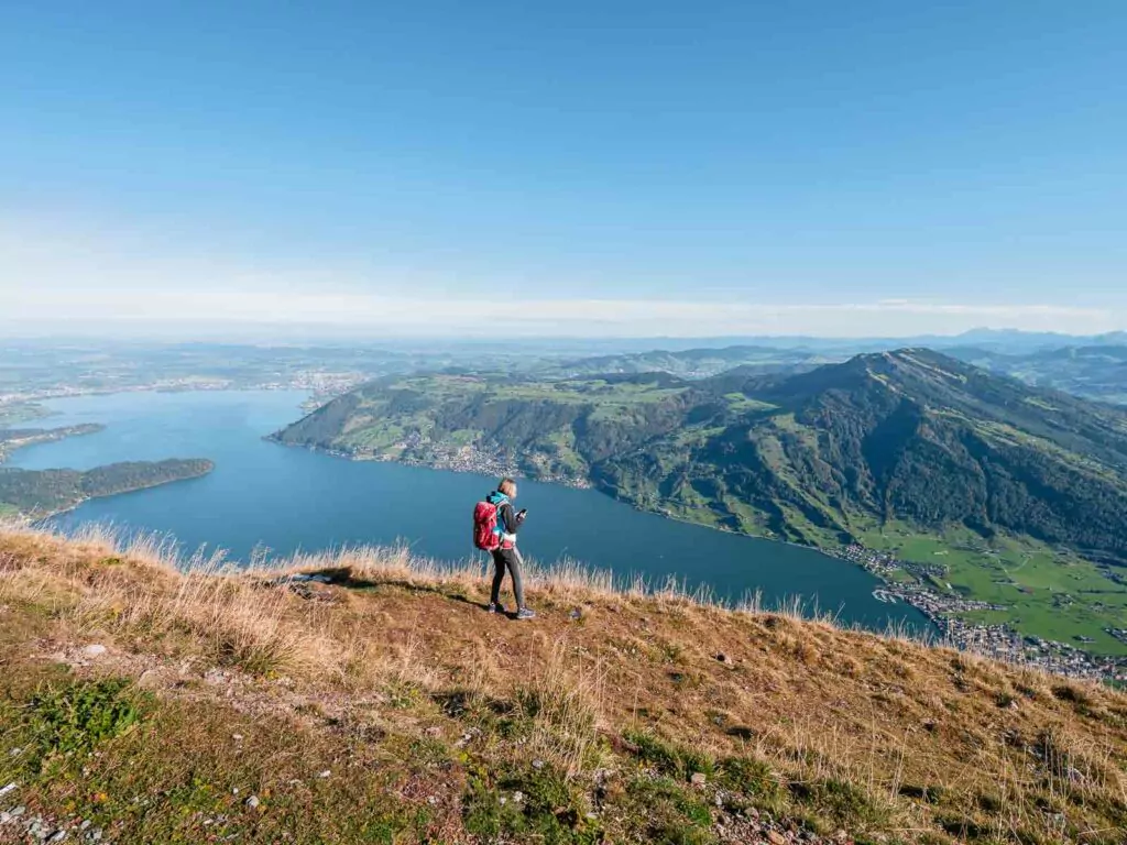 A hiker standing on a grassy slope of Mount Rigi, overlooking the expansive view of a lake and surrounding mountains under a clear sky