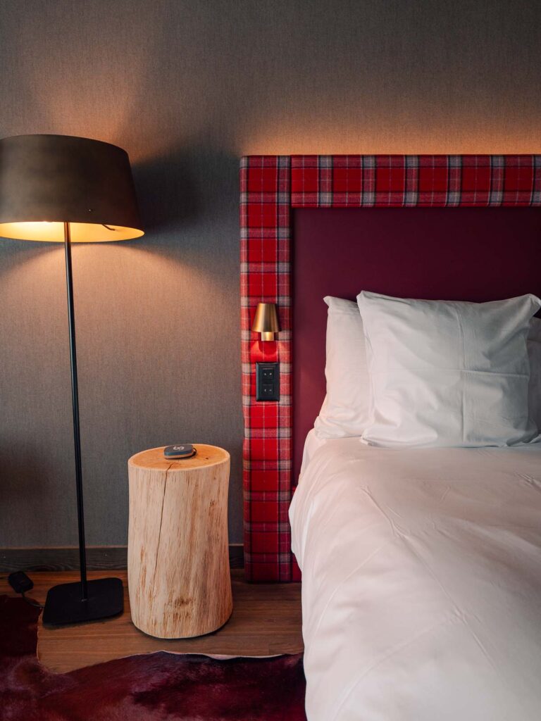 Hotel room at Bergwelt Grindelwald with a red plaid headboard, a white pillow on a bed, a wooden bedside table, and a floor lamp