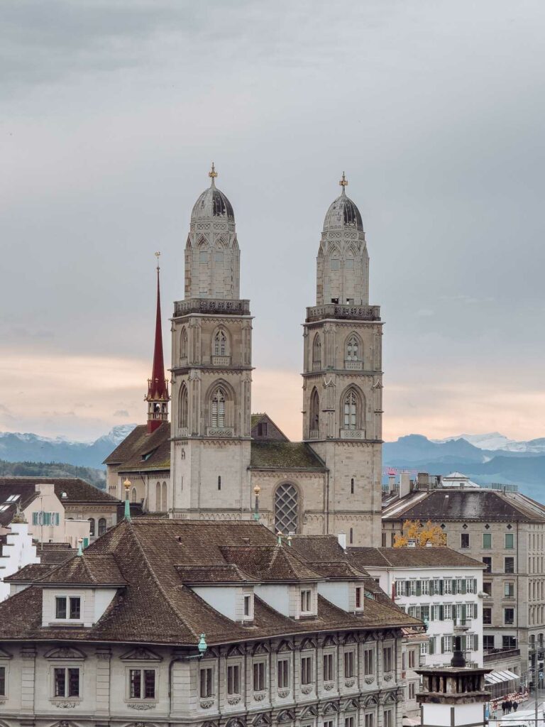 The Grossmünster church in Zurich, showcasing its twin towers against a backdrop of the city skyline and distant mountains under a cloudy sky