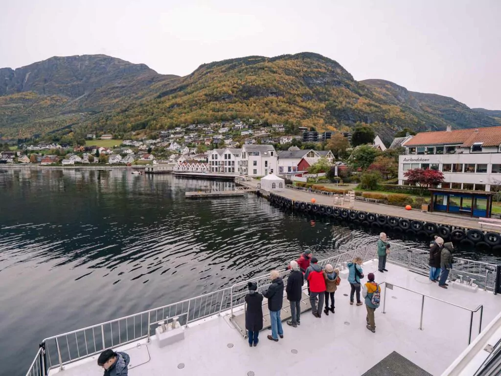 Tourists on the deck of a Naeroyfjord cruise observe the scenic waterfront and undulating hills of a Norwegian village, a captivating experience for a three-week Scandinavia itinerary