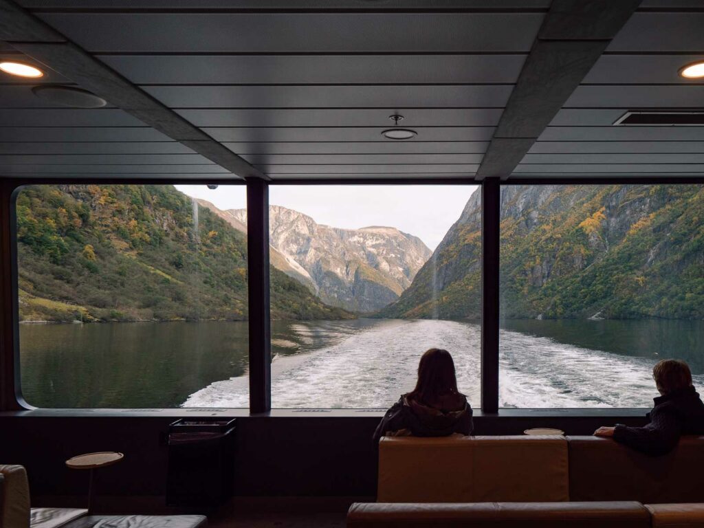 Silhouetted figures inside the Naeroyfjord cruise boat lounge gaze out of the panoramic windows at the receding fjord, its calm waters lined by steep verdant cliffs, capturing the peaceful retreat into nature's embrace