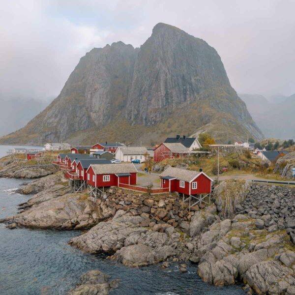 The iconic red rorbuer cabins of Hamnøy in the Lofoten Islands, with a majestic mountain backdrop and clear waters in the foreground, a quintessential stop on a Scandinavia itinerary
