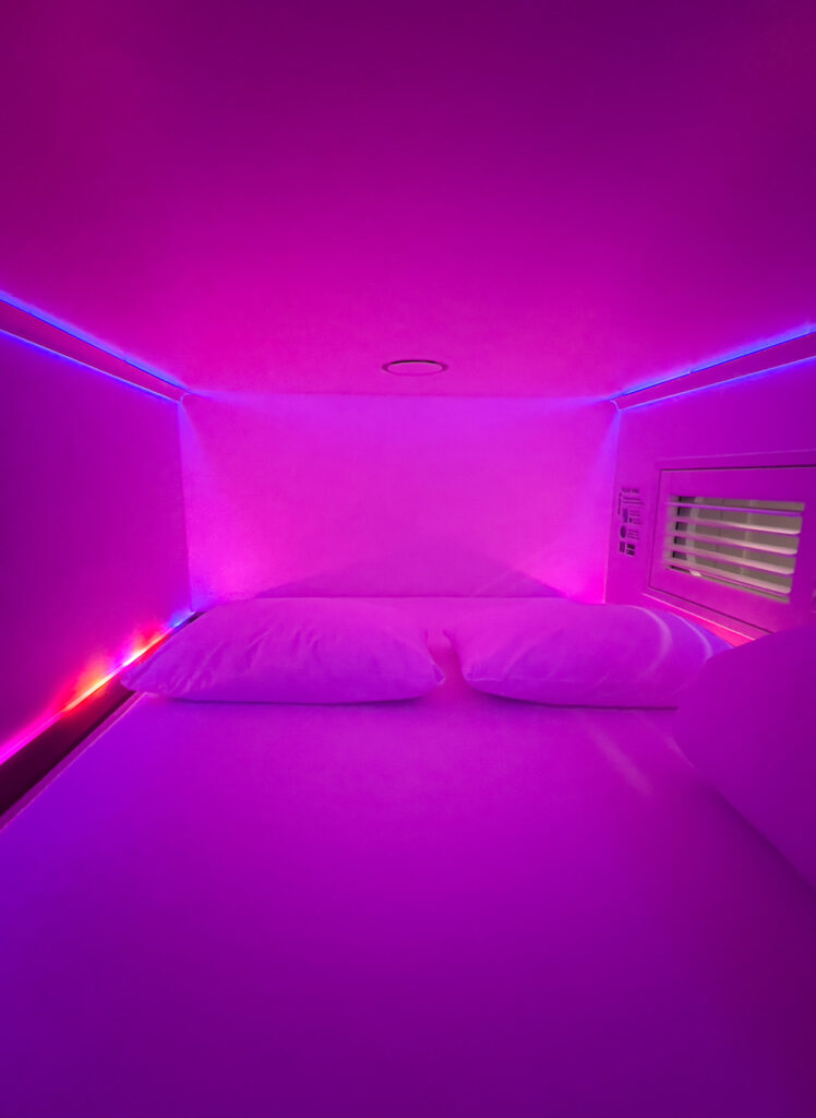 Inside a CityHub Copenhagen sleeping pod, bathed in vibrant pink light, with a cozy white bed offering a unique and modern accommodation experience