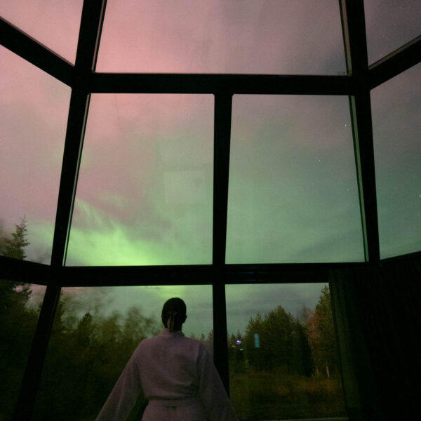 A silhouetted figure stands in awe inside Santa's Igloos Arctic Circle in Rovaniemi, gazing out at the ethereal Northern Lights dancing across the night sky through the igloo's glass ceiling