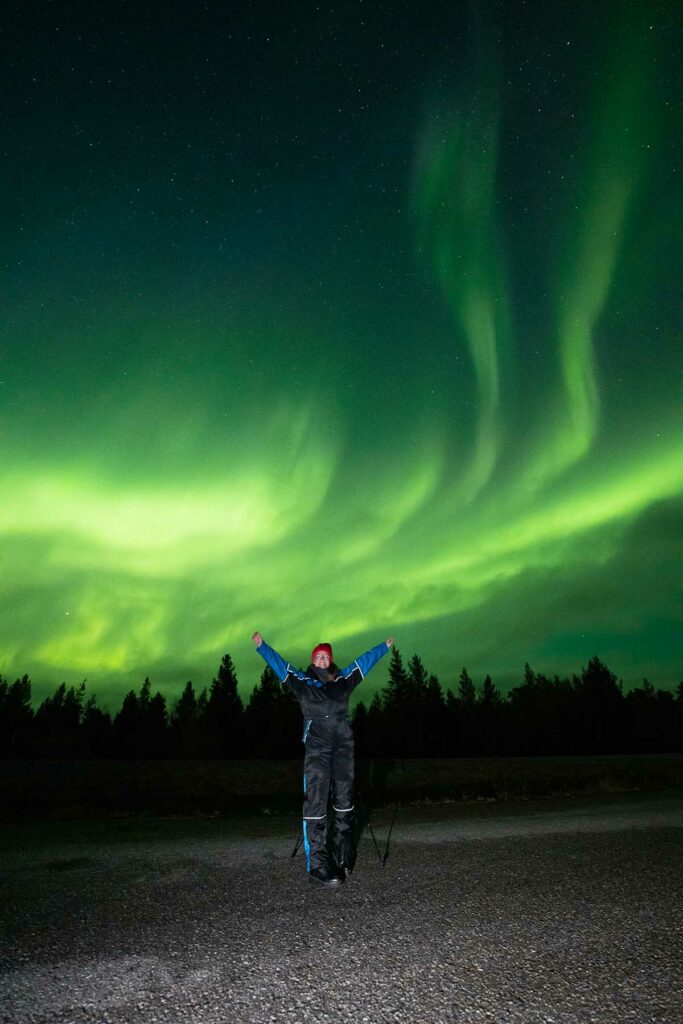 A person named Alexx standing with arms raised under the aurora borealis in Rovaniemi, the vibrant green Northern Lights creating a spectacular backdrop in the Finnish sky
