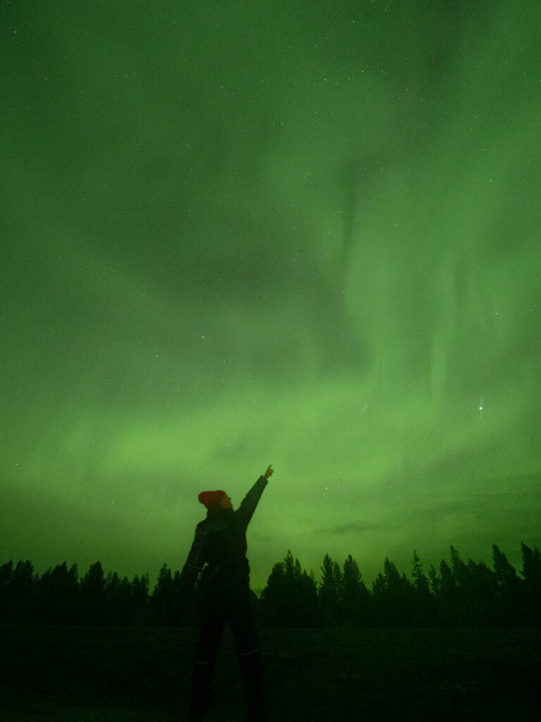A person named Alexx standing with arms raised under the aurora borealis in Rovaniemi, the vibrant green Northern Lights creating a spectacular backdrop in the Finnish sky