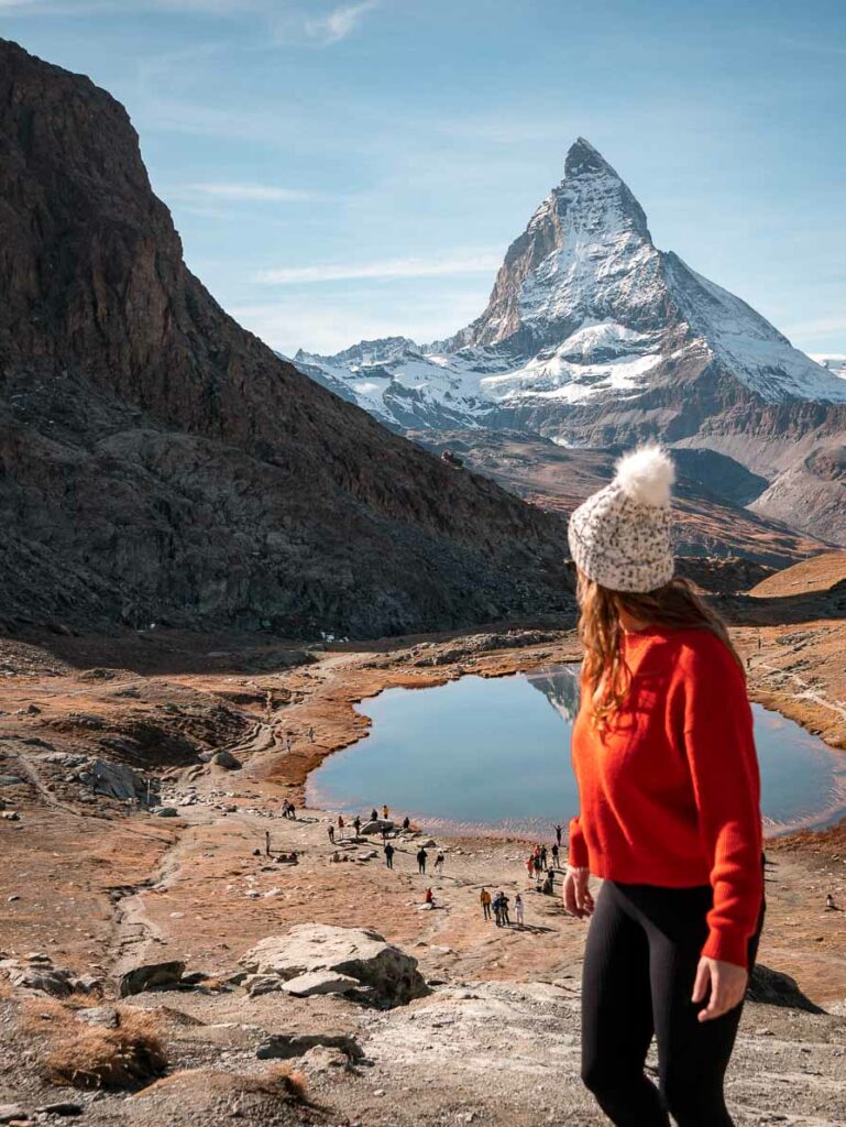 Alexx in a red sweater and white beanie standing before the Matterhorn and Riffelsee lake with other visitors