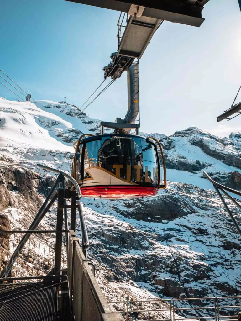 The Titlis Rotair cable car with its distinctive red and yellow colors, approaching the station with a snowy mountainous landscape in the background