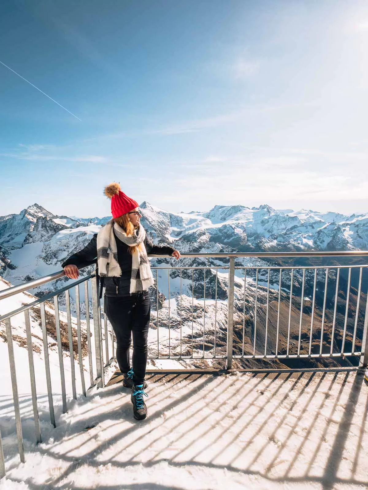 A person standing on a snow-covered viewing platform at the summit of Mount Titlis, overlooking a vast expanse of snow-capped mountains under a clear blue sky
