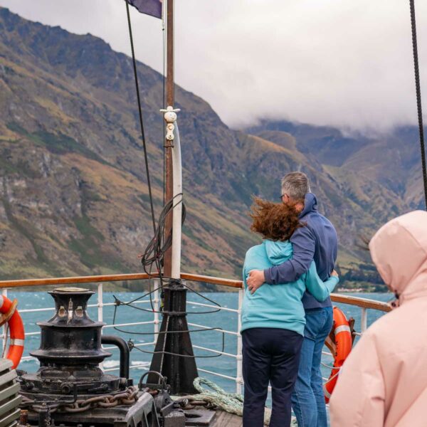 Passengers on the deck of the TSS Earnslaw steamship, with a couple embracing and looking out at the mountainous landscape around Lake Wakatipu, New Zealand