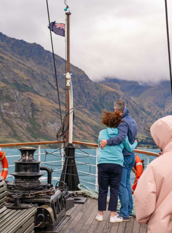 Passengers on the deck of the TSS Earnslaw steamship, with a couple embracing and looking out at the mountainous landscape around Lake Wakatipu, New Zealand