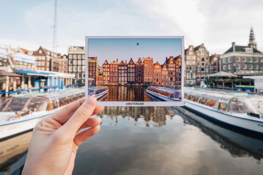 Creative composition with a postcard of traditional Amsterdam canal houses held in the foreground, perfectly aligning with the actual scene in the background.