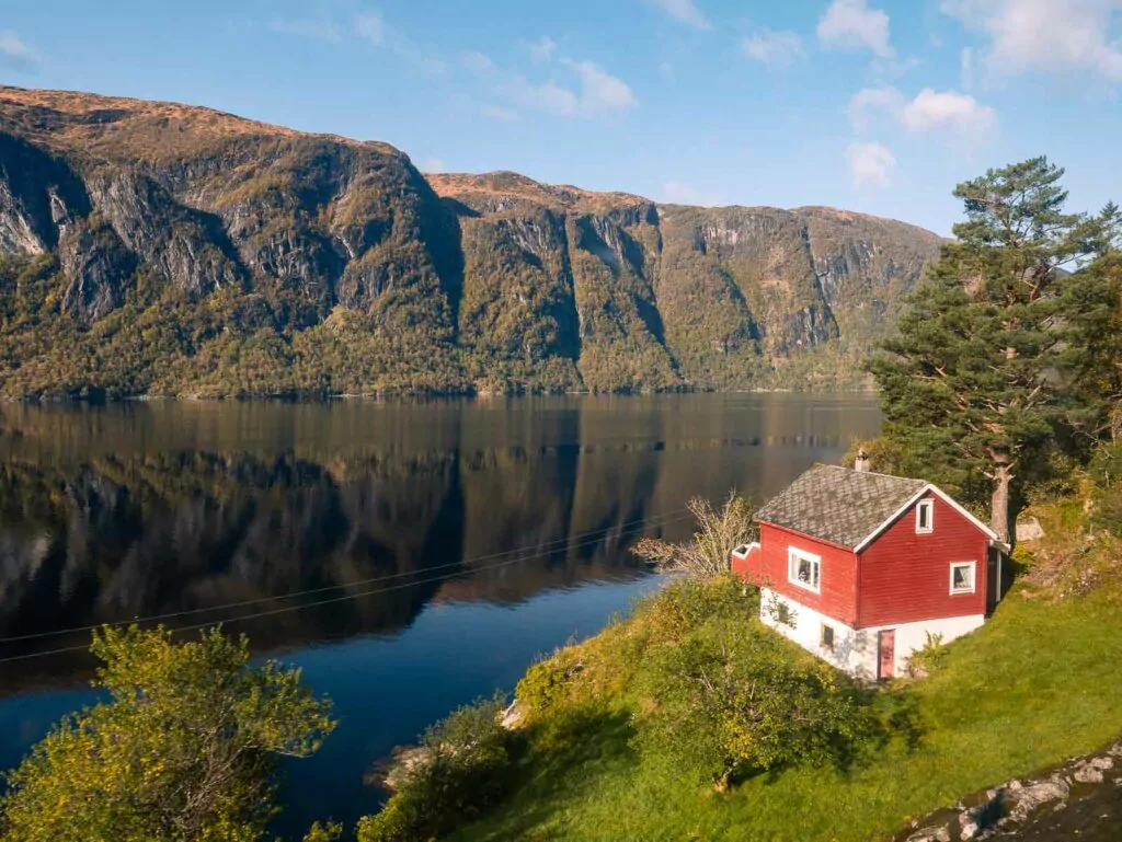 A solitary red house stands on the edge of a tranquil lake, reflecting the clear skies and mountainous backdrop, a scene visible from the Bergen Railway Line in Norway