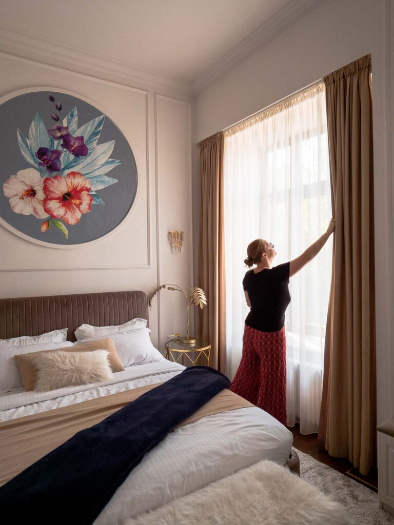 A woman opens the curtains in a sunlit hotel room in Bucharest, revealing a large floral wall art, offering a glimpse into the elegant accommodations for a 2 weeks Balkans itinerary