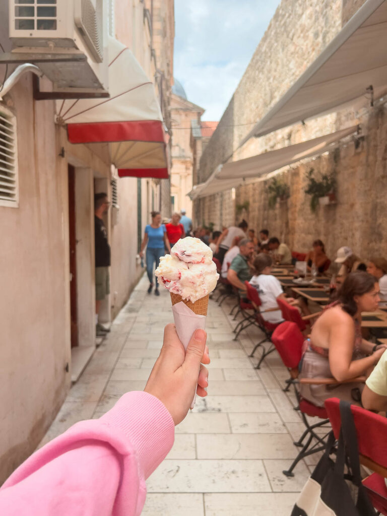 First-person perspective of a strawberry ice cream cone held up against a narrow street lined with outdoor café seating in Dubrovnik