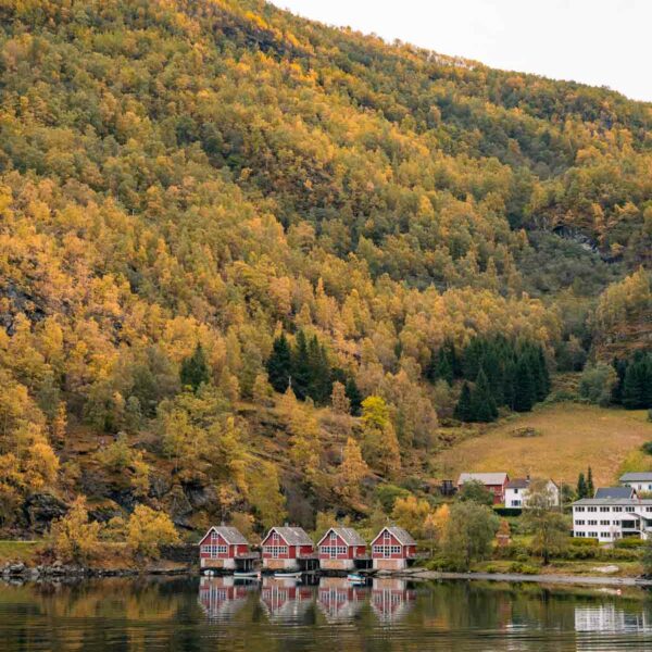 The calm waters of Aurlandsfjord, Norway, reflect a row of quaint red and white boathouses at the base of a forested hillside bathed in the soft light of dusk