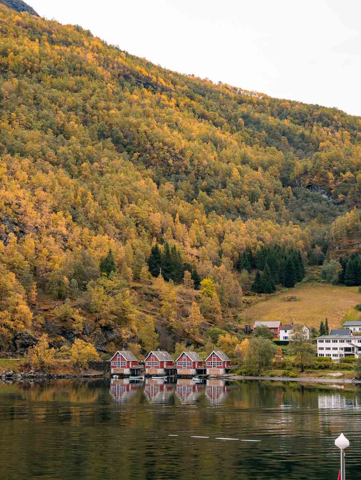 The calm waters of Aurlandsfjord, Norway, reflect a row of quaint red and white boathouses at the base of a forested hillside bathed in the soft light of dusk