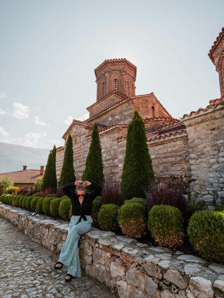 A woman in a blue dress leans on a stone wall along a cobblestone path near the St. John at Kaneo Church, with perfectly trimmed green hedges, at sunset in Ohrid, Macedonia