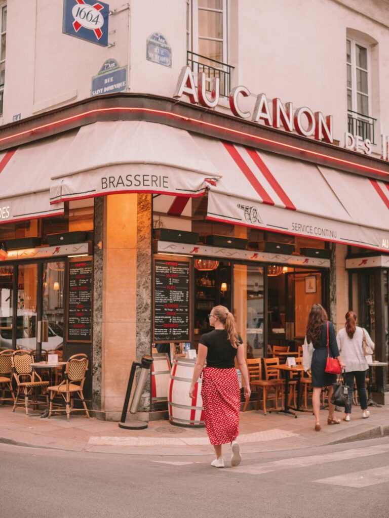 A woman walks past the quaint entrance to the Au Canon Des Invalides brasserie in Paris, with its classic Parisian charm inviting passersby.