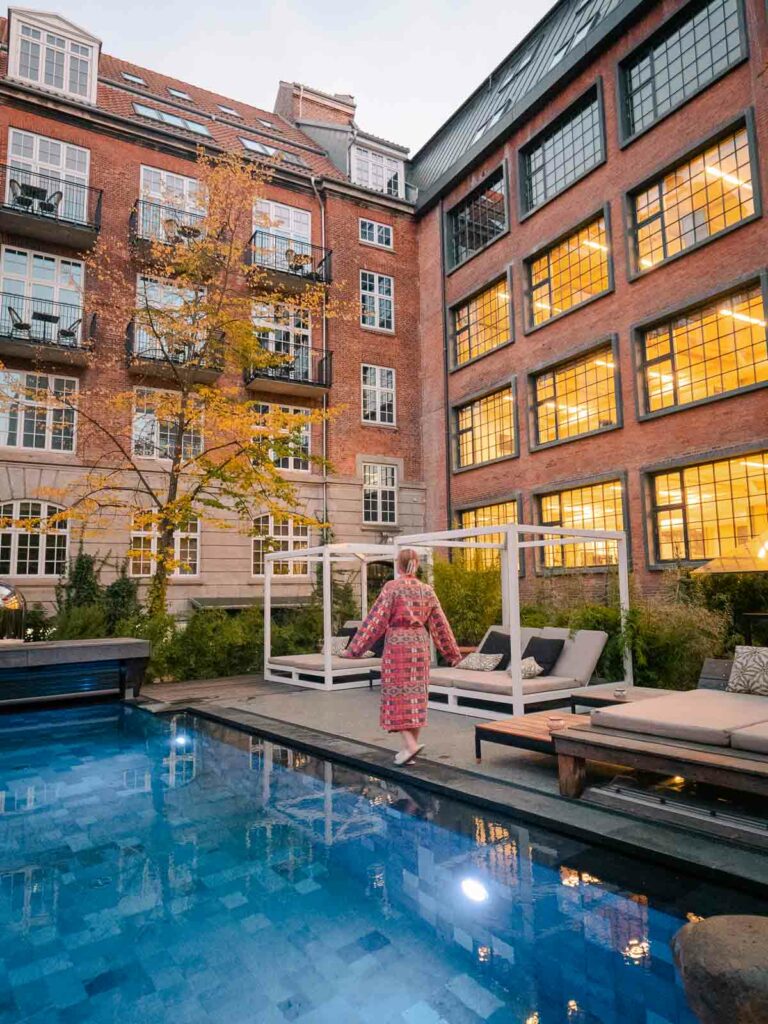 Alexx in a patterned dress stands by a serene outdoor pool, flanked by traditional Danish architecture, capturing the essence of relaxation while staying at Bryggen Guldsmeden on a solo trip to Copenhagen