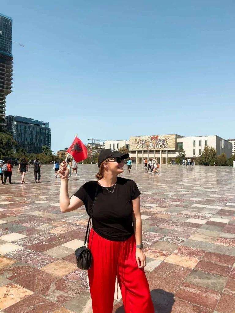 Woman in a black top and red trousers standing in Skanderbeg Square, Tirana, with the National History Museum in the background