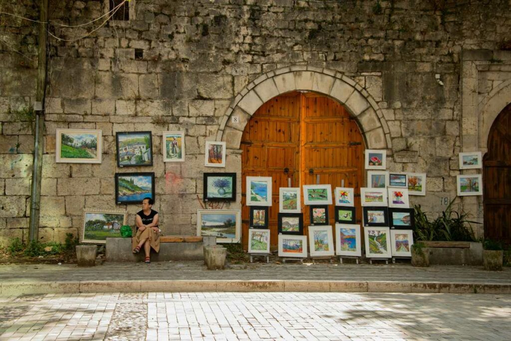 An artist's display of colourful paintings set against an old stone wall with a wooden door in the Albanian Riviera, showcasing local art