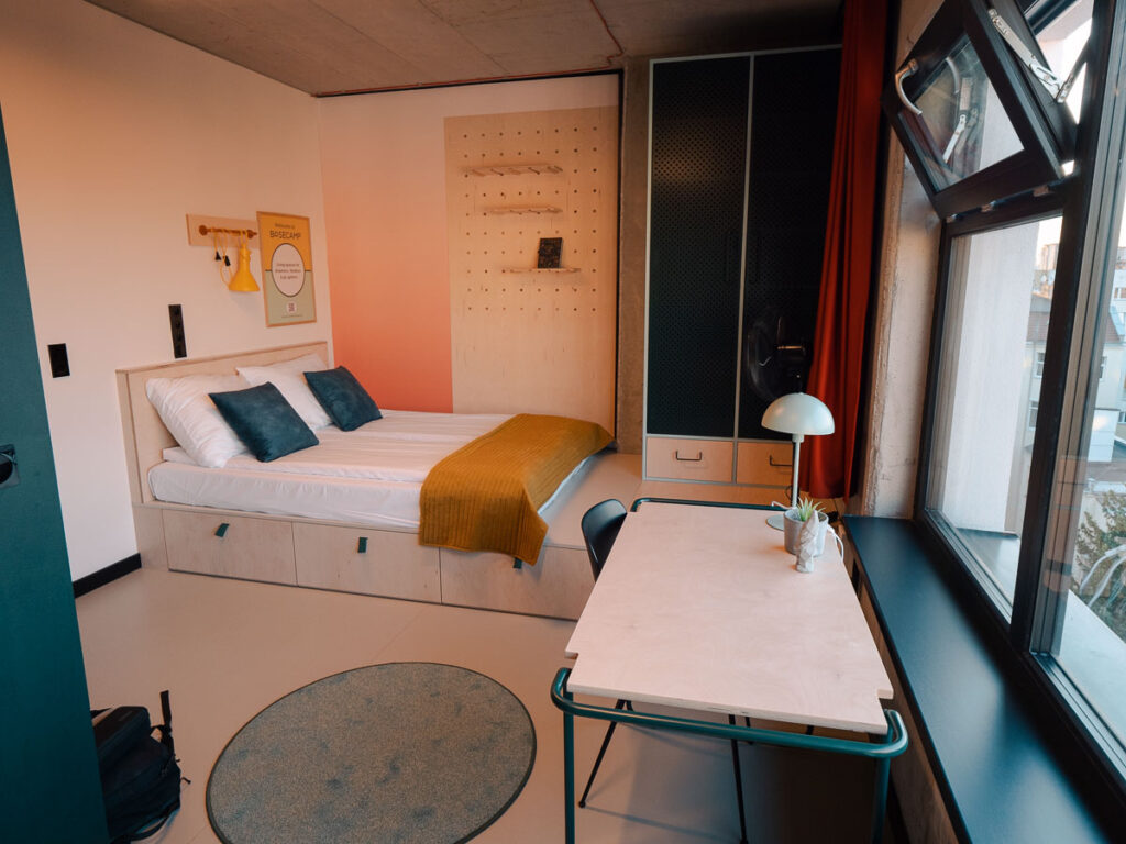 A cozy and modern studio apartment at Basecamp in Wroclaw, featuring a well-lit work area and a snug bed, framed by distinctive geometric windows.