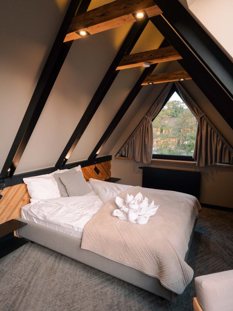 A cozy attic bedroom at Villa T in Zakopane, featuring a plush bed with grey and white bedding, nestled under wooden beams with warm lighting and a view of the trees through angular windows