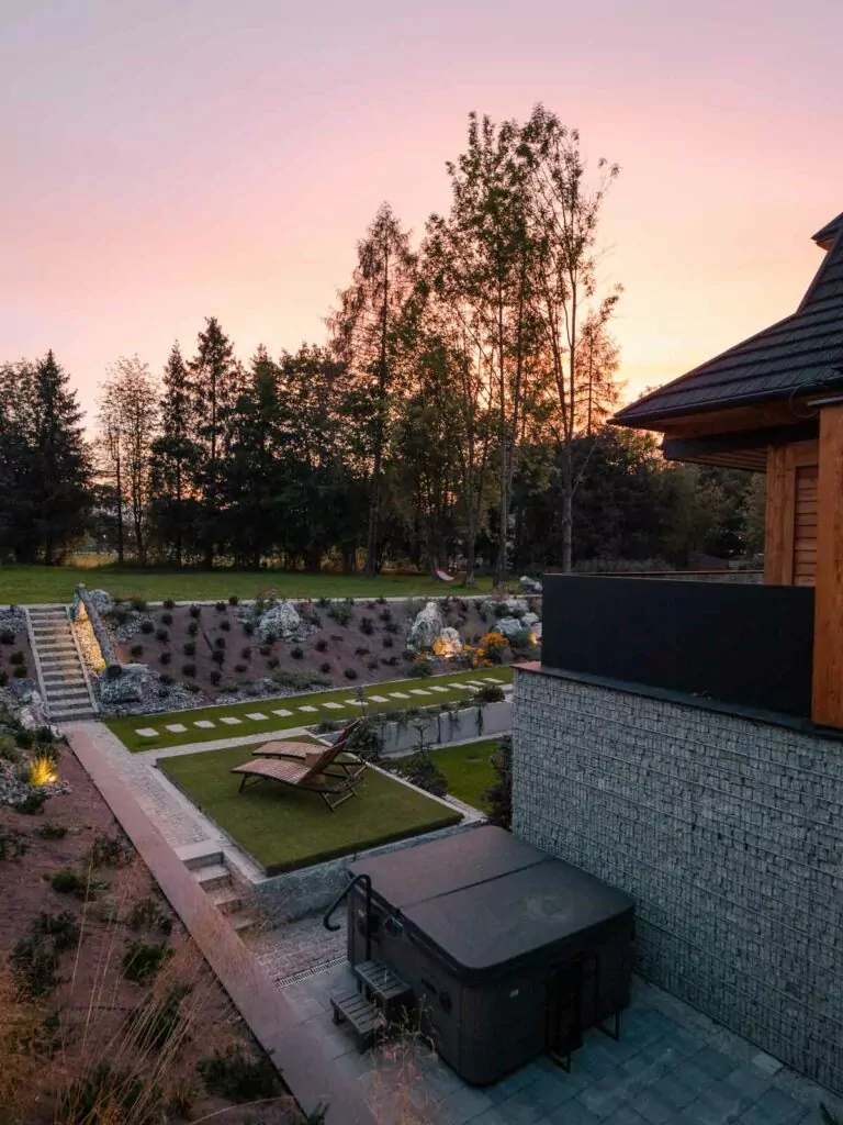 The tranquil backyard of Villa T in Zakopane during twilight, featuring a modern hot tub and a neatly landscaped garden, inviting relaxation amid the encircling trees