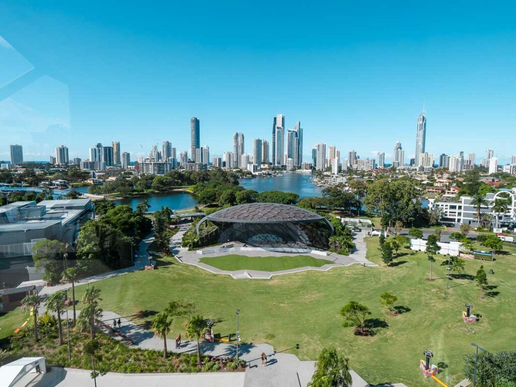 A sprawling green park featuring a bandstand, with the impressive city skyline of the Gold Coast in the background, providing a serene oasis within the urban landscape