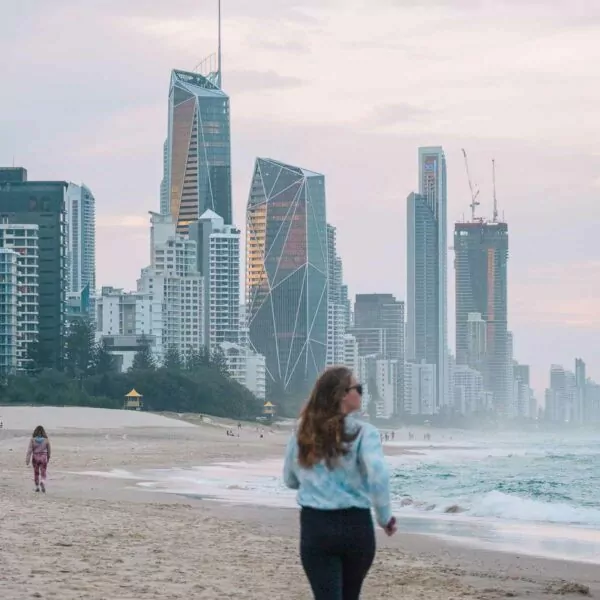 A woman strolling along the beach at sunset with the Gold Coast city skyline in the background, creating a picturesque scene of urban and natural beauty
