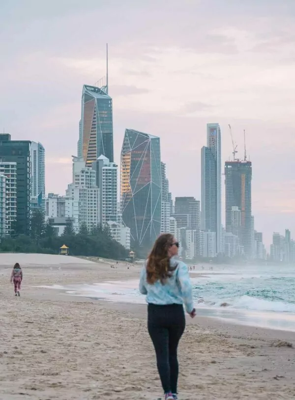 A woman strolling along the beach at sunset with the Gold Coast city skyline in the background, creating a picturesque scene of urban and natural beauty