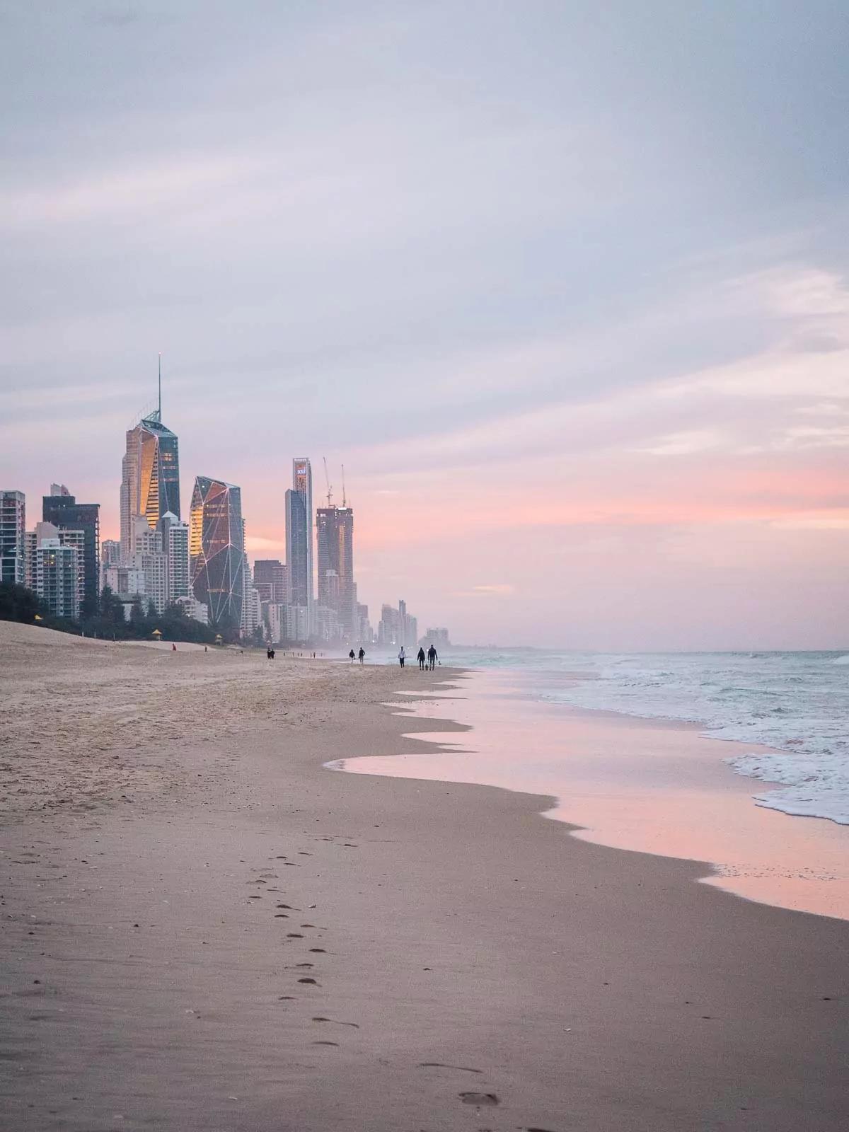 A serene beach with gentle waves washing up on the shore, footprints in the sand leading towards a distant city skyline illuminated by the soft hues of sunset, capturing the beauty of the Gold Coast